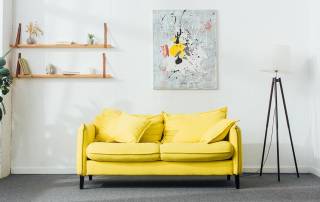 Ein Zimmer mit einem gelben Sofa, einer Lampe, Pflanzen sowie einem Bild und einem Regal an der Wand | Home Staging