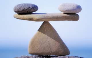 Eine Waage aus Steinen im Gleichgewicht am Meer, ein dreieckiger Stein steht auf dem Boden, auf seiner Spitze liegt ein flacher Stein, auf dessen Enden rechts und links jeweils ein Stein liegt, alles ist im Gleichgewicht | Scheidung Zugewinngemeinschaft Güterstand