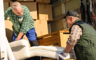 Zwei Männer heben in einem Umzugswagen voller Kisten ein Sofa an - Wohnen im Alter