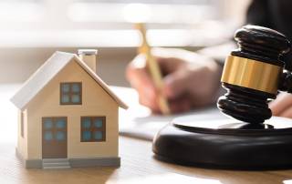 Ein Mann unterschreibt ein Dokument auf einem Tisch und im Vordergrund stehen ein Modellhaus und ein Richterhammer | Immobilie in der Scheidung