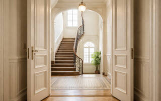 Helle, Altbau-Eigentumswohnung in einem gepflegten Mehrfamilienhaus, Blick vom Flur der Wohnung durch eine offene Altbau-Wohnungstür in ein sauberes, Altbau-Treppenhaus, viel natürliches Tageslicht, aufgeräumt und hochwertig, professionelle Immobilienfotografie, realistisch, hohe Auflösung, weiche Lichtstimmung | Immobilienverkauf