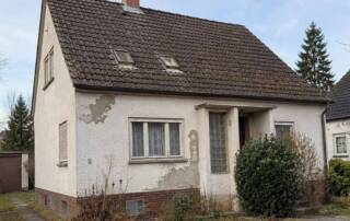 Ein älteres Einfamilienhaus mit verwitterter Fassade und sanierungsbedürftigem Dach steht in einer Wohnstraße und symbolisiert die Entscheidung zwischen Modernisierung und Verkauf. I Sanierungspflicht beim Verkaufen